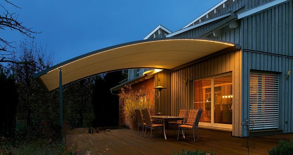 Eine gebogene Markise mit unterseitig angebrachten Lichtleisten erhellen Sitzgelegenheiten auf einer Terrasse.